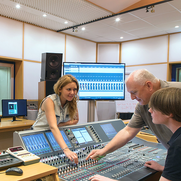 In a recording studio, two students and a teacher stand at a mixing desk.