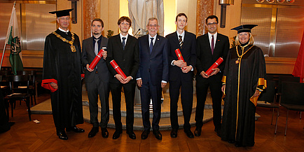 Eine Gruppe Menschen in festlichem Gewand steht in der Aula der TU Graz. Die vier Männer in der Mitte halten rote Diplomrollen in Händen.