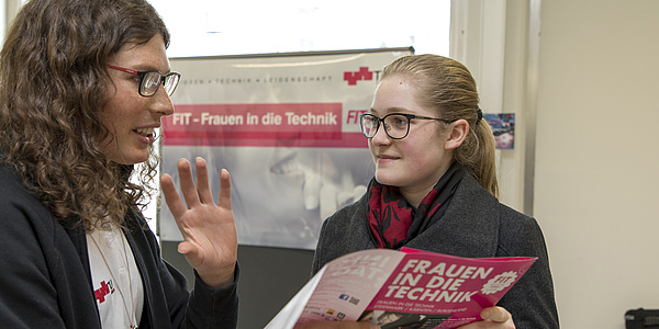 2 Frauen unterhalten sich. Eine Frau hält eine Broschüre in der Hand.