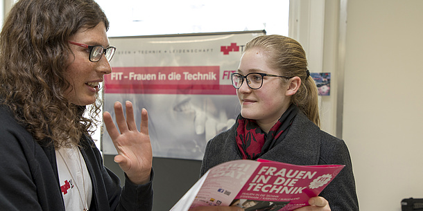 2 Frauen unterhalten sich. Eine Frau hält eine Broschüre in der Hand.