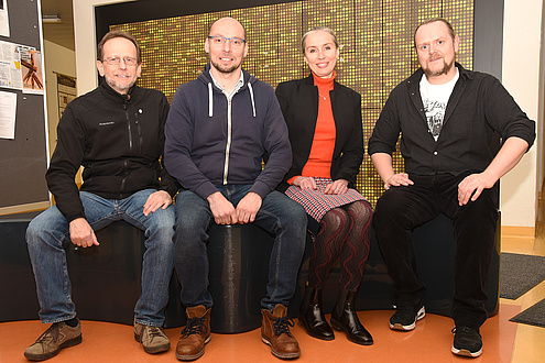 Gruppenfoto von drei Männern und einer Frau, alle nebeneinander sitzend