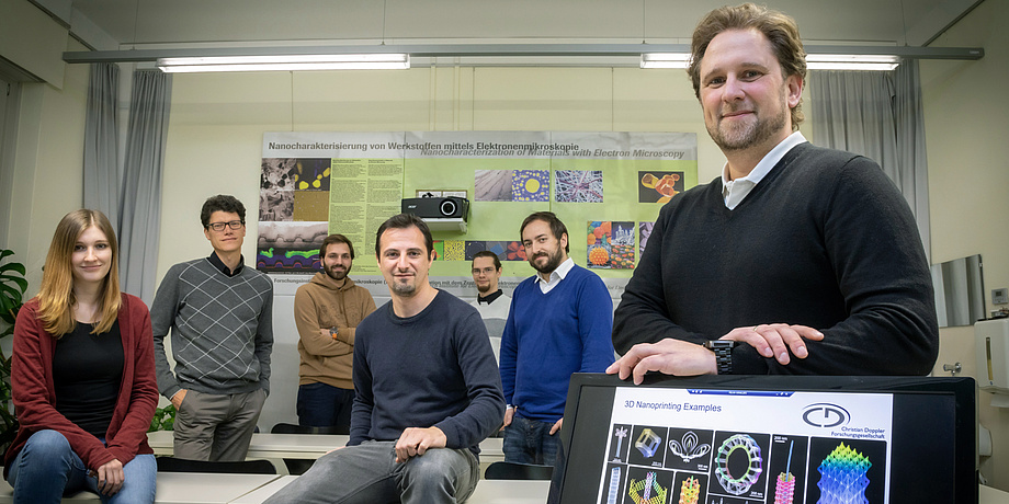 Gruppe von Menschen in Büro, rechts Bildschirm mit Abbildungen von Nanostrukturen, hinten ein Poster zu Nanoanalytik