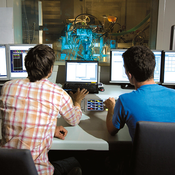 Two men in front of several screens, a machine is working behind a glass wall.