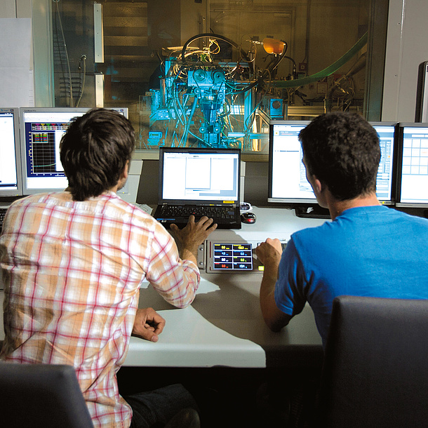 Two men in front of several screens, a machine is working behind a glass wall. 