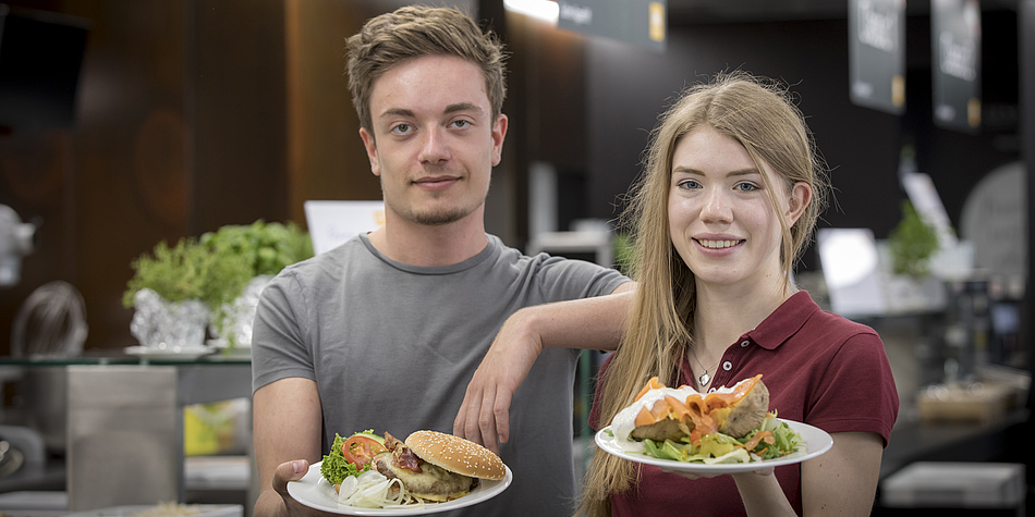 Two people are at the canteens counter. A young man dressed in a grey shirt and a young woman in red. Both are holding plates of food.