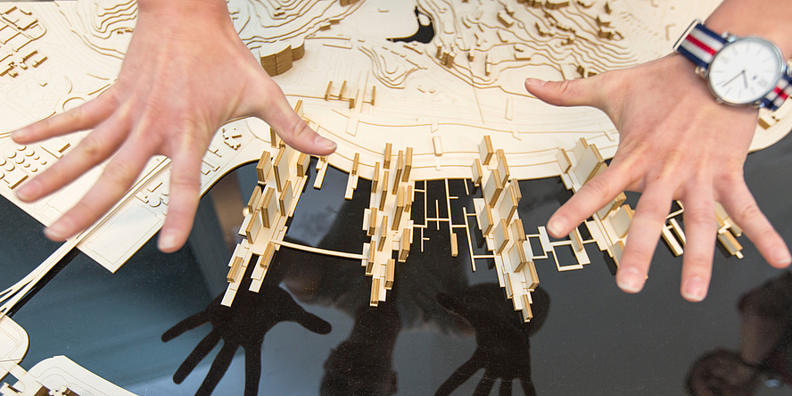 Two hands spread out over an architectural model made of wood.