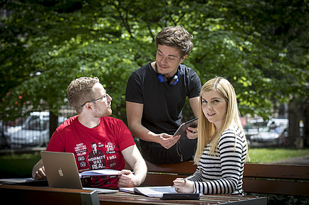 Drei junge Leute, ein Mann im roten T-Shirt daneben ein Mann im schwarzen T-Shirt und eine junge Frau mit blonden Haaren sitzen in der Natur und tauschen sich aus.