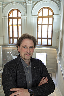 A man in a grey shirt smiles, standing in front of two large windows.