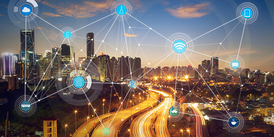 Eine beleuchtete Straße und Gebäude im Hintergrund. Davor ist ein Netzwerk zu sehen mit vielen kleinen Symbolen, die unter anderem Computer, W-LAN-Symbole und Autos darstellen.