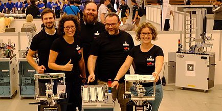 Three men and two women in black TU Graz T-shirts pose together with two robots in a production hall