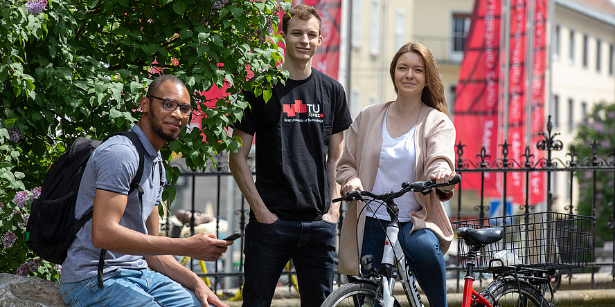 Ein Student mit schwarzem Rucksack, sitzend, ein Student mit TU Graz-Shirt, stehend und eine Studentin am Fahrrad vor dem schmiedeisernen Zaun und roten TU Graz-Fahnen am Gelände der Alten Technik.