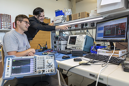 Zwei Männer an einem Tisch mit mehreren elektronischen Geräten, vielen Kabeln und kleinen Bauteilen.