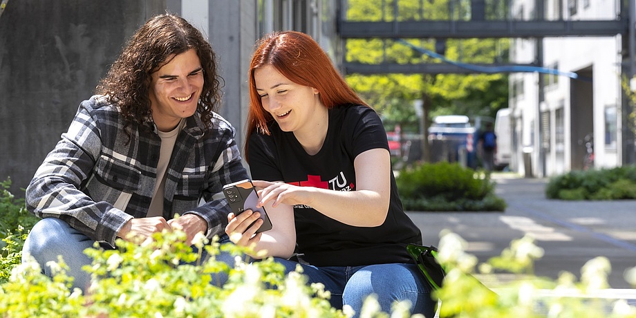 Eine Studentin und ein Student sitzen am Campus, unterhalten sich und schauen dabei auf ein Handy.