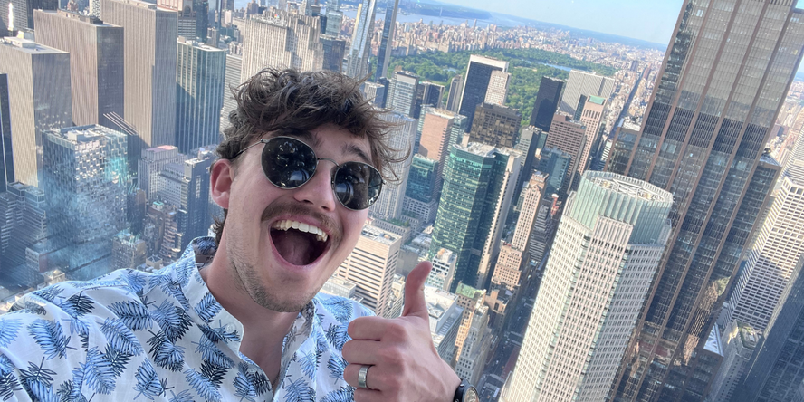 Ein junger Mann grinst mit erhobenen Daumen in die Kamera, unten hinter ihm die Skyline von New York.