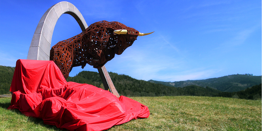 TU Graz Racing Team (Mit roter Plane verhüllter Bolide auf einer grünen Wiese vor der mehr als lebensgroßen Stahlskulptur eines Stiers unter einem über 17 m hohen Stahlbogen.