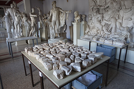 Die Fragmente einer Altarplatte liegen auf einem Tisch, im Hintergrund antike Statuen.