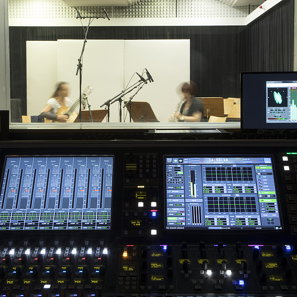 Recording studio with mixing desk. Behind a disc two musicians with instruments sit in the recording room.