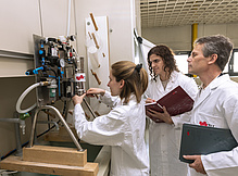 Eine Frau hantiert an einem Laborgerät, zwei Männer schauen interessiert zu.