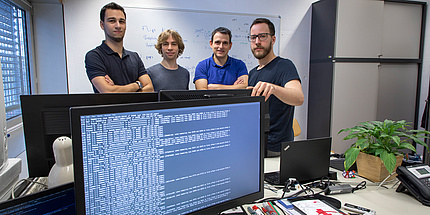 Ein großer Computerbildschirm mit vielen Zeichen ist im Vordergrund zu sehen. Dahinter stehen vier Männer.