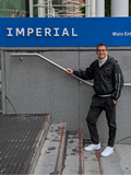 A smiling man in black sportswear with white sneakers leans on a handrail in front of gray steps beneath a blue sign reading “IMPERIAL.”