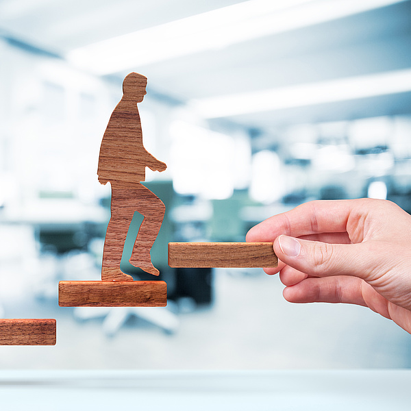 Eine Hand hält eine flache Holzstufe, die das Ende einer Stiege darstellt. Eine Holzfigur steigt die Stufen hinauf. Im Hintergrund ist ein Büro zu sehen.