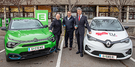 Drei lächelnde Männer stehen zwischen zwei Elektroautos, zwei davon halten ein Ladekabel in der Hand.