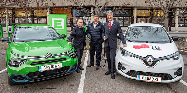 Three smiling men stand between to electric vehicles, two of them hold a charging cable