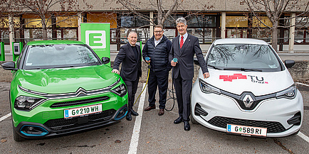 Drei lächelnde Männer stehen zwischen zwei Elektroautos, zwei davon halten ein Ladekabel in der Hand.