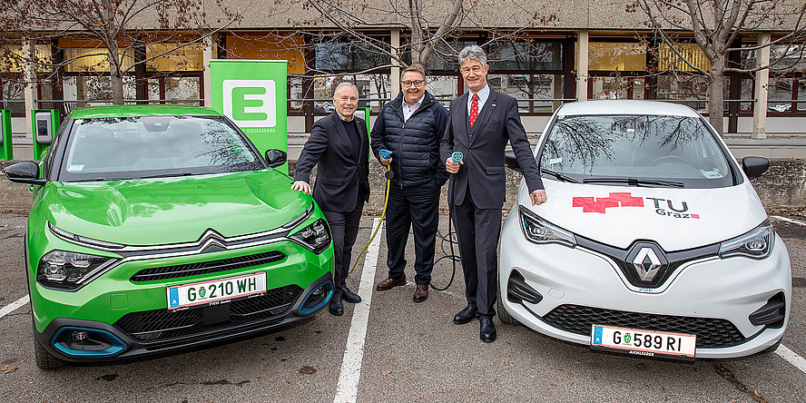 Three smiling men stand between to electric vehicles, two of them hold a charging cable
