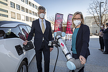 Ein Mann und eine Frau, beide einen Mundschutz (Maske) tragend, tanken gerade ein E-Auto. 