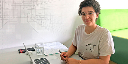 Junge Frau sitzt an einem Schreibtisch mit Laptop, Schreibunterlagen, Buch und Wasserglas.
