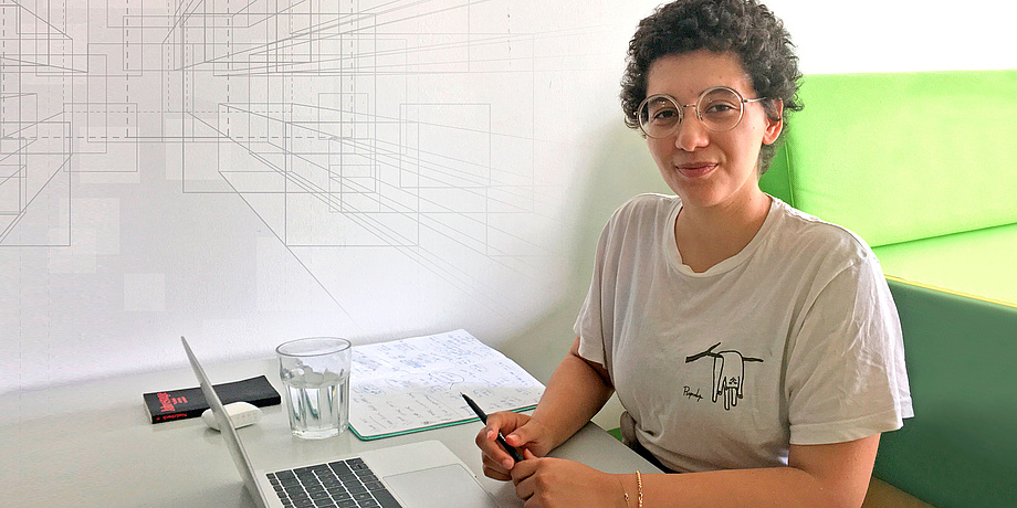 Young woman sitting at a desk with laptop, writing pads, book and water glass.