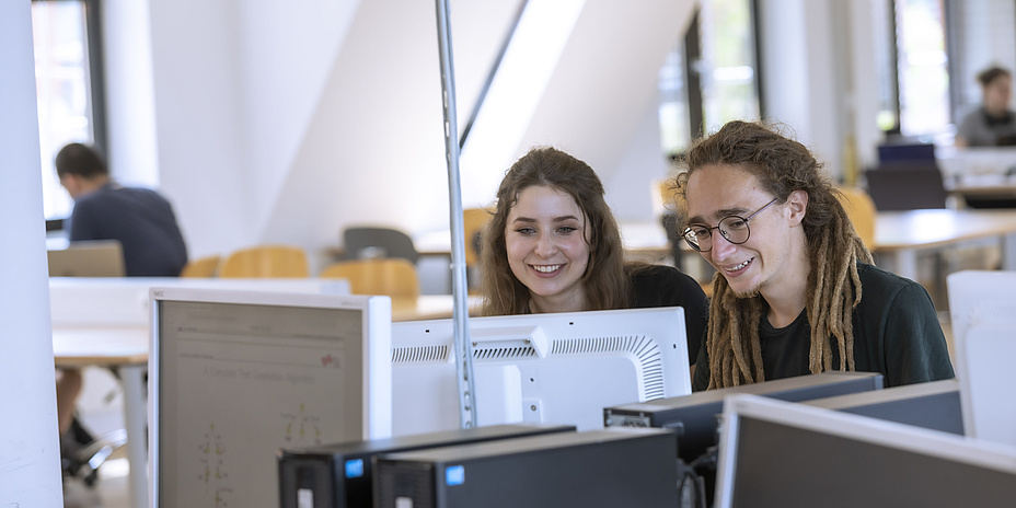 A young woman and a young man work together on a PC in a bright room with many computer workstations.