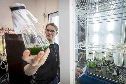 A woman stands in front of a kind of refrigerator and holds a glass flask with green liquid in the camera.