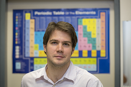 Young TU Graz researcher stands in front of a periodic table poster