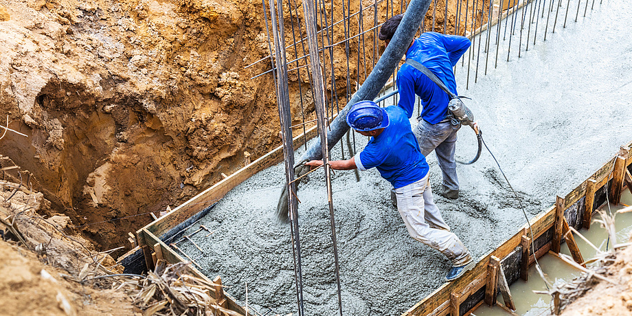 Two construction workers stand in a wooden enclosure and spread concrete that comes out of a hose.