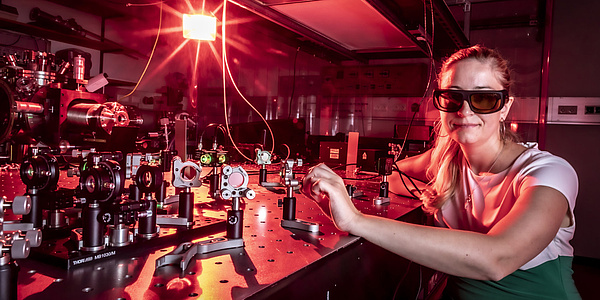 A woman is sitting in a laboratory next to a table with an experimental setup, wearing safety goggles.