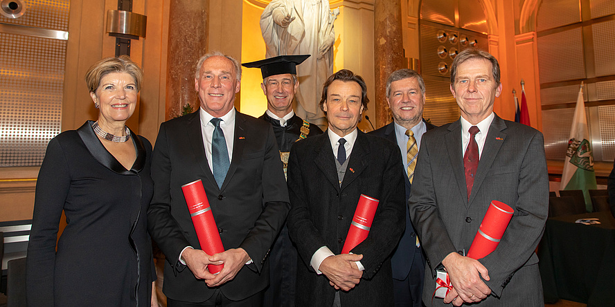Eine Frau und fünf Männer stehen nebeneinander in festlicher Kleidung, drei Männer halten eine rote Urkundenrolle in Händen