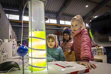 Drei Kinder blicken auf eine Röhre mit fluoriszierender Flüssigkeit.