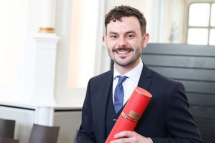 A man holds up a red document roll and smiles into the camera.