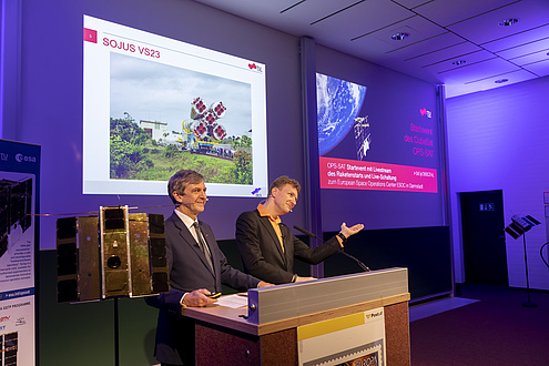 Zwei Männer sprechen vor Publikum; im Hintergrund Powerpoint Folien