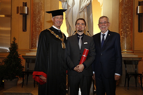 Drei Männer in festlichem Gewand, jener in der Mitte hält eine rote Diplomrolle in der Hand.