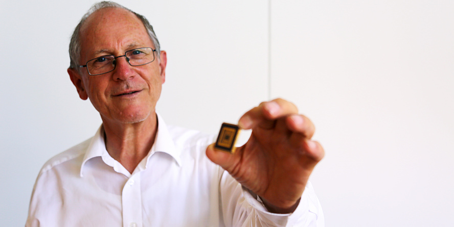 A men is holding a black, square plate with golden lines on it. He has grey hair and is wearing a white shirt.