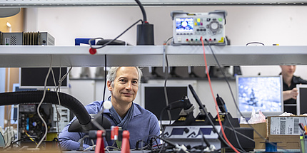 A man sits between technical devices and smiles into the camera