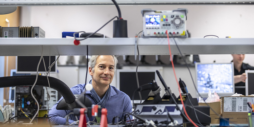 A man sits between technical devices and smiles into the camera