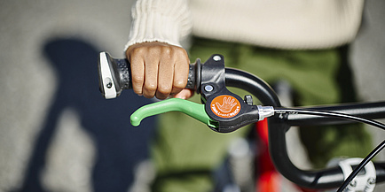 A child's hand grips a bicycle handlebar. In front of it is a green brake lever