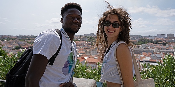 Zwei Personen lächeln in die Kamera vor einem Panoramaausblick über Lissabon.