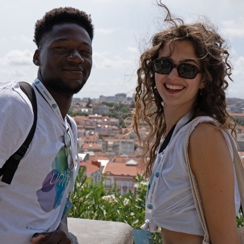 Zwei Personen lächeln in die Kamera vor einem Panoramaausblick über Lissabon.
