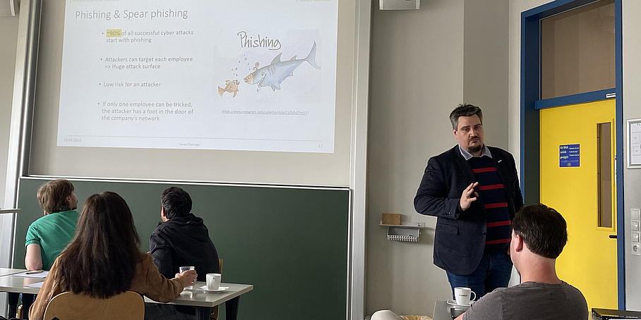 Lecturer in a teaching situation with 4 students explains a projected presentation slide on the topic "Phishing & Spear Phishing"