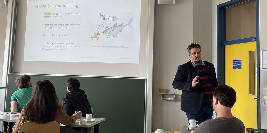 Vortragender in einer Unterrichtssituation mit 4 Studierenden erklärt eine projizierte Präsentationsfolie zum Thema "Phishing & Spear Phishing"